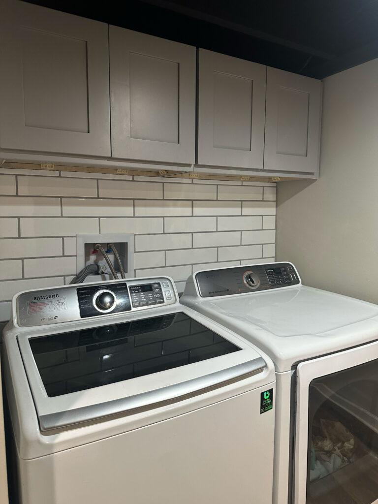 Laundry area with gray shaker upper cabinets and a white subway tile backsplash above a washer and dryer