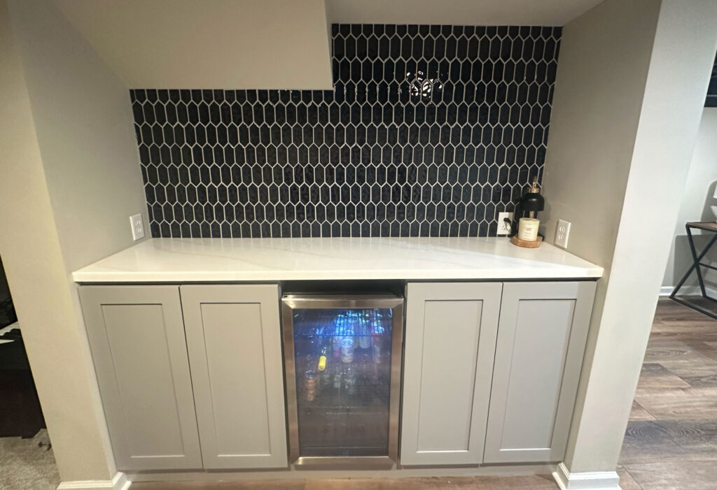 Basement bar area with black elongated hexagon tile backsplash, white countertop, gray shaker cabinets and a beverage fridge